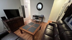 Un salón con un sofá de cuero marrón oscuro, una mesa de centro de madera, un televisor sobre un mueble de madera y un espejo redondo en la pared.
