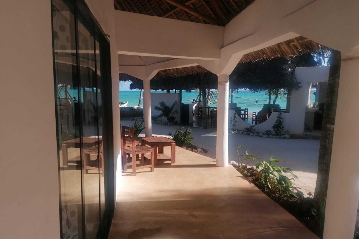 Un espace extérieur aménagé en terrasse, avec vue sur la plage, des tables et des chaises en bois, et des structures dotées de toits en chaume.