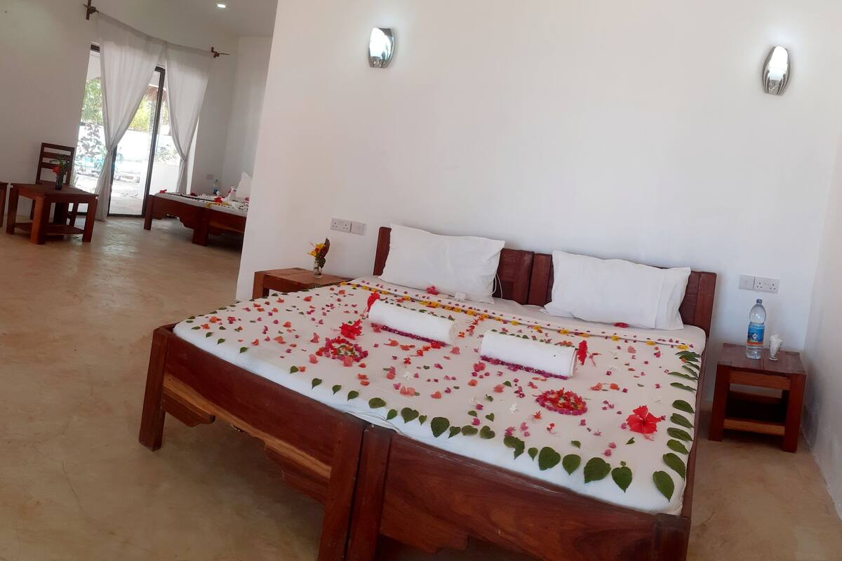 Une chambre d’hôtel avec un grand lit, une tête de lit en bois, du linge de lit blanc et une petite table de chevet avec une bouteille d’eau.
