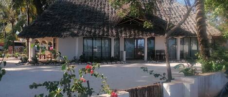 A thatched-roof building with large windows, surrounded by palm trees and a garden with pink flowers.