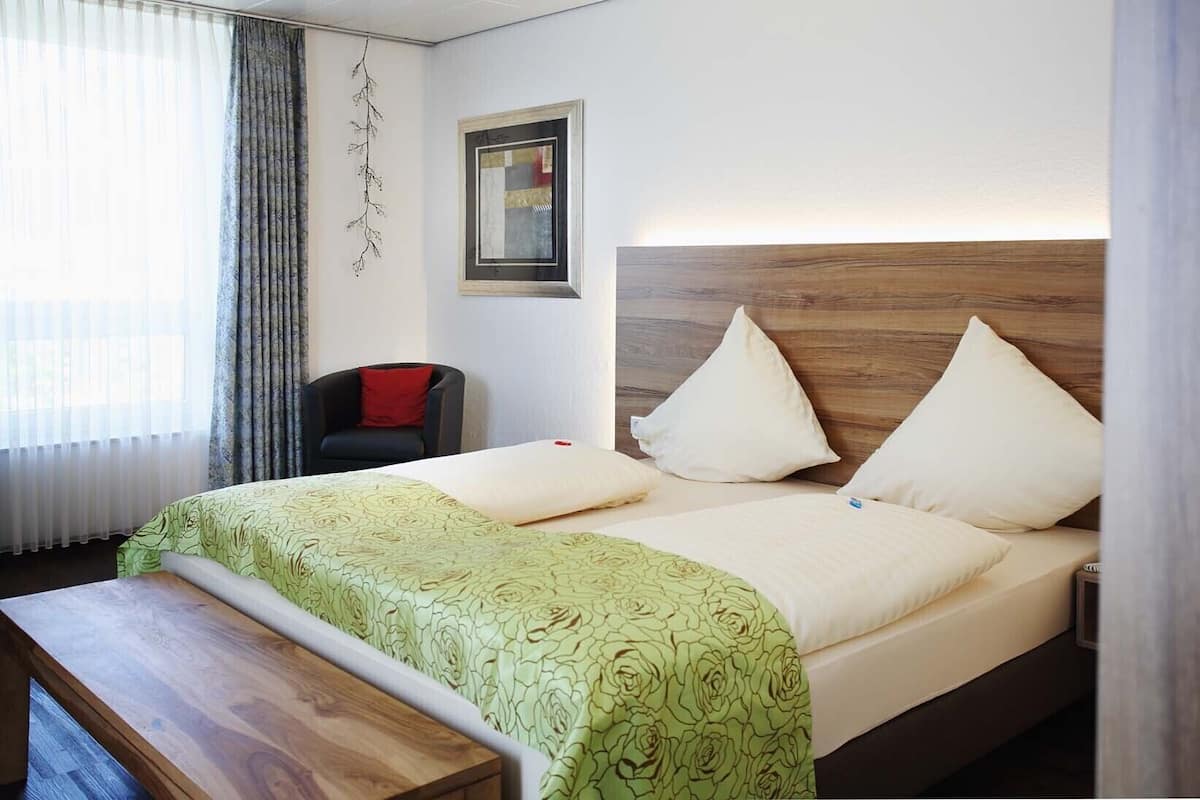 A hotel room with a bed, a bench, a chair, and a window with curtains.