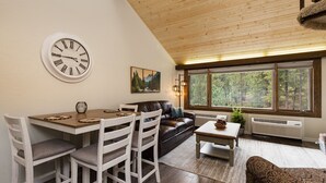 En hyggelig stue med spiseplads, med træloft, store vinduer med udsigt til en skov og et vægur.