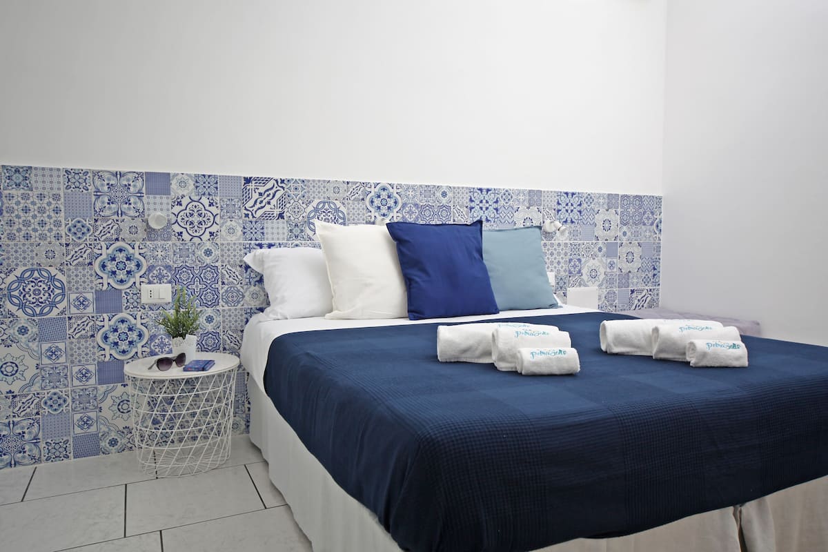 Una cama bien hecha con colcha y almohadas azules, una mesita de noche blanca con una planta y gafas de sol, y una pared con estampado azul y blanco.