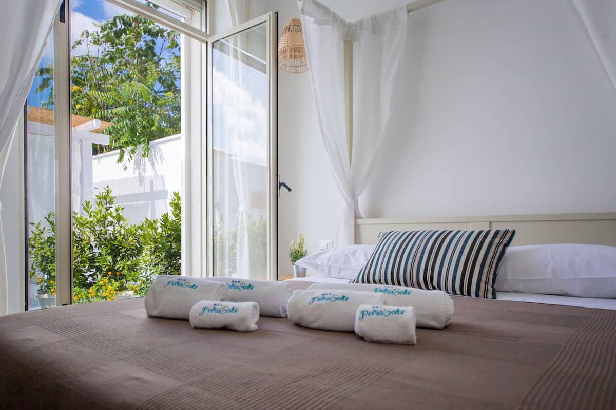 Habitación de hotel con cama, cortinas blancas, vistas a zonas verdes y toallas con texto.