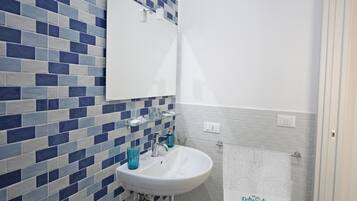 Un baño con un lavamanos blanco, paredes con azulejos azules y grises, un estante de vidrio con artículos de aseo y una toalla en un perchero.
