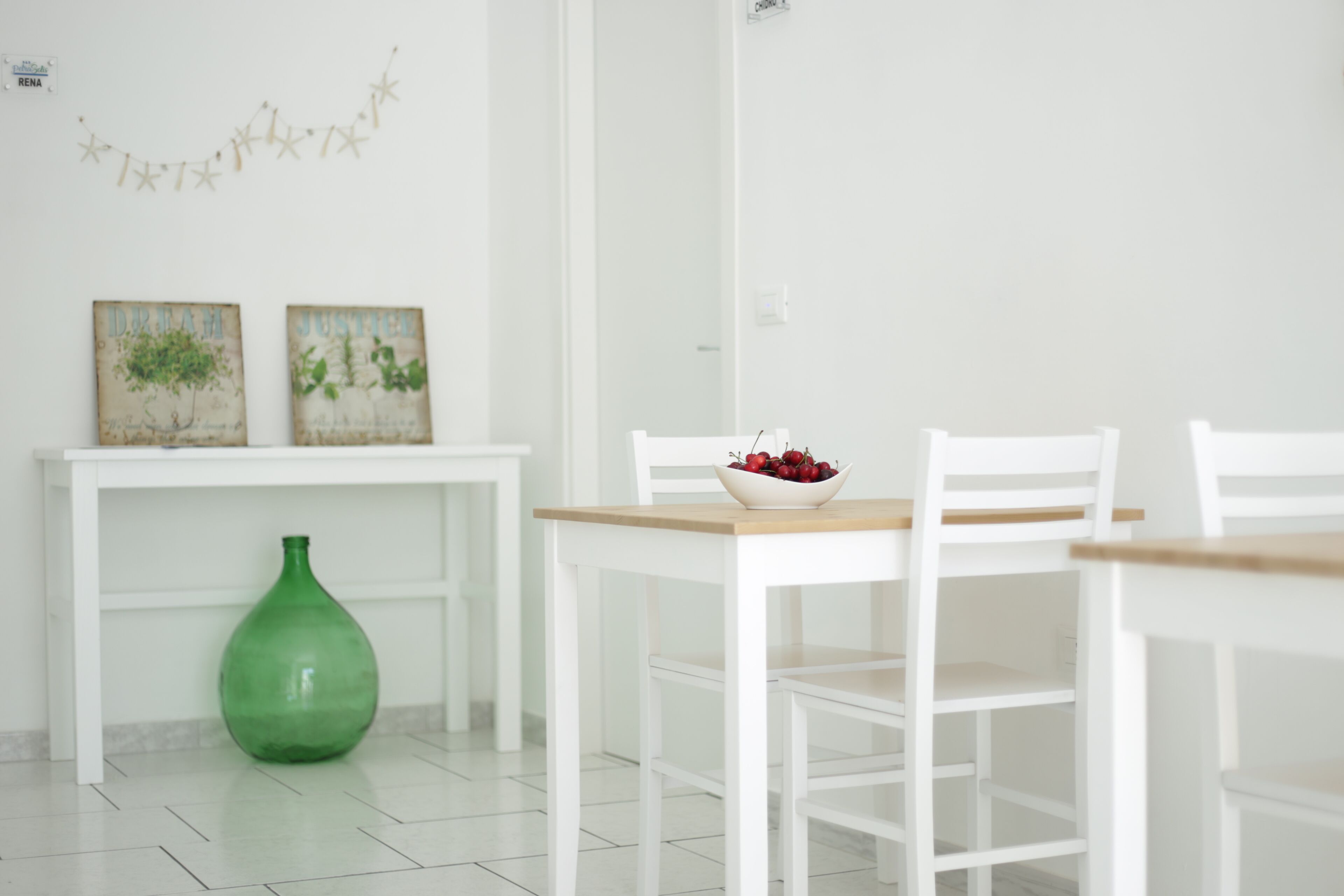 A white dining area with a table, chairs, a bowl of fruit, and two framed pictures on a white wall.