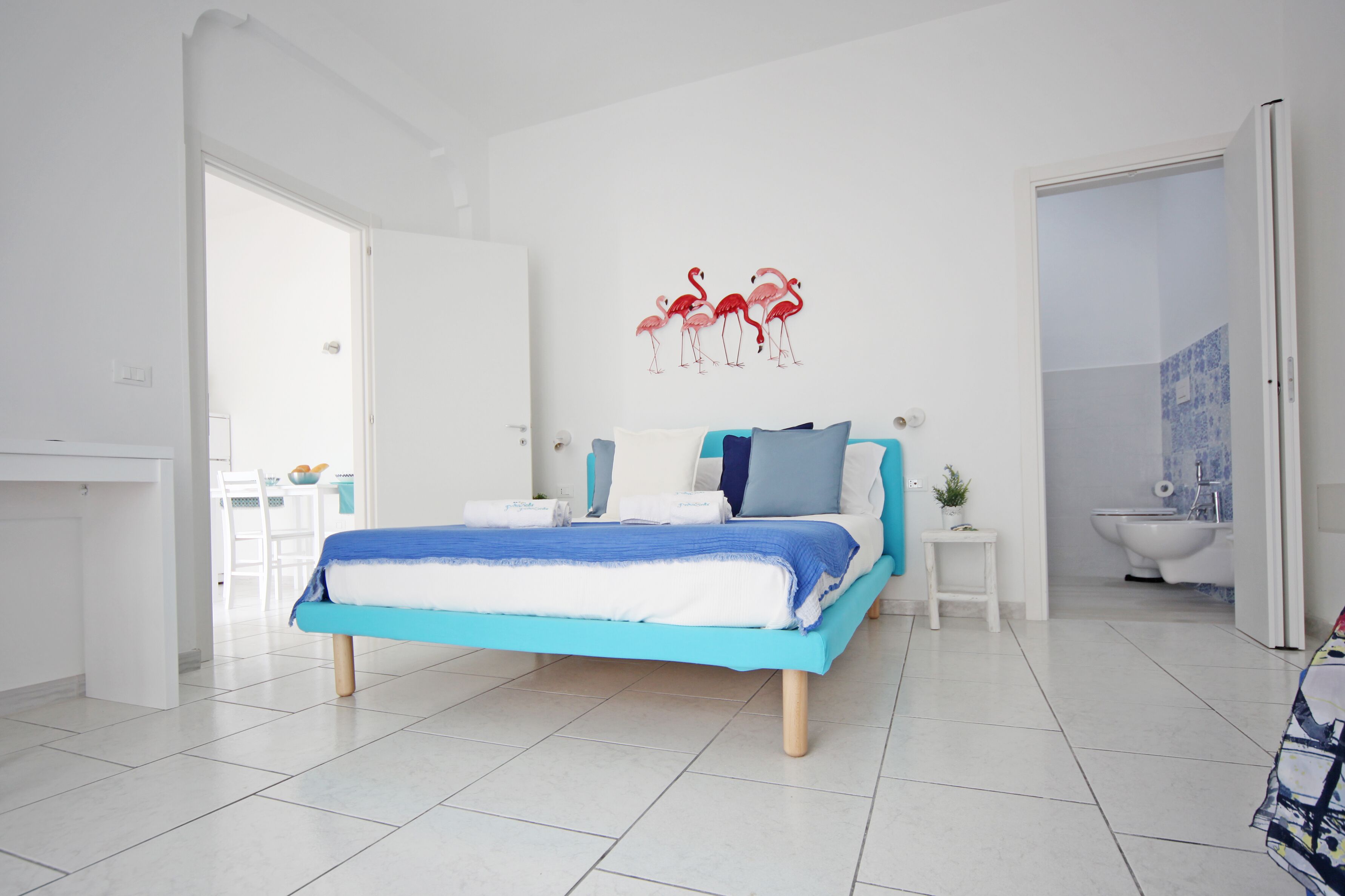 A modern bedroom with a bed, a white door, a bathroom with a toilet, and a wall with a flamingo decoration.