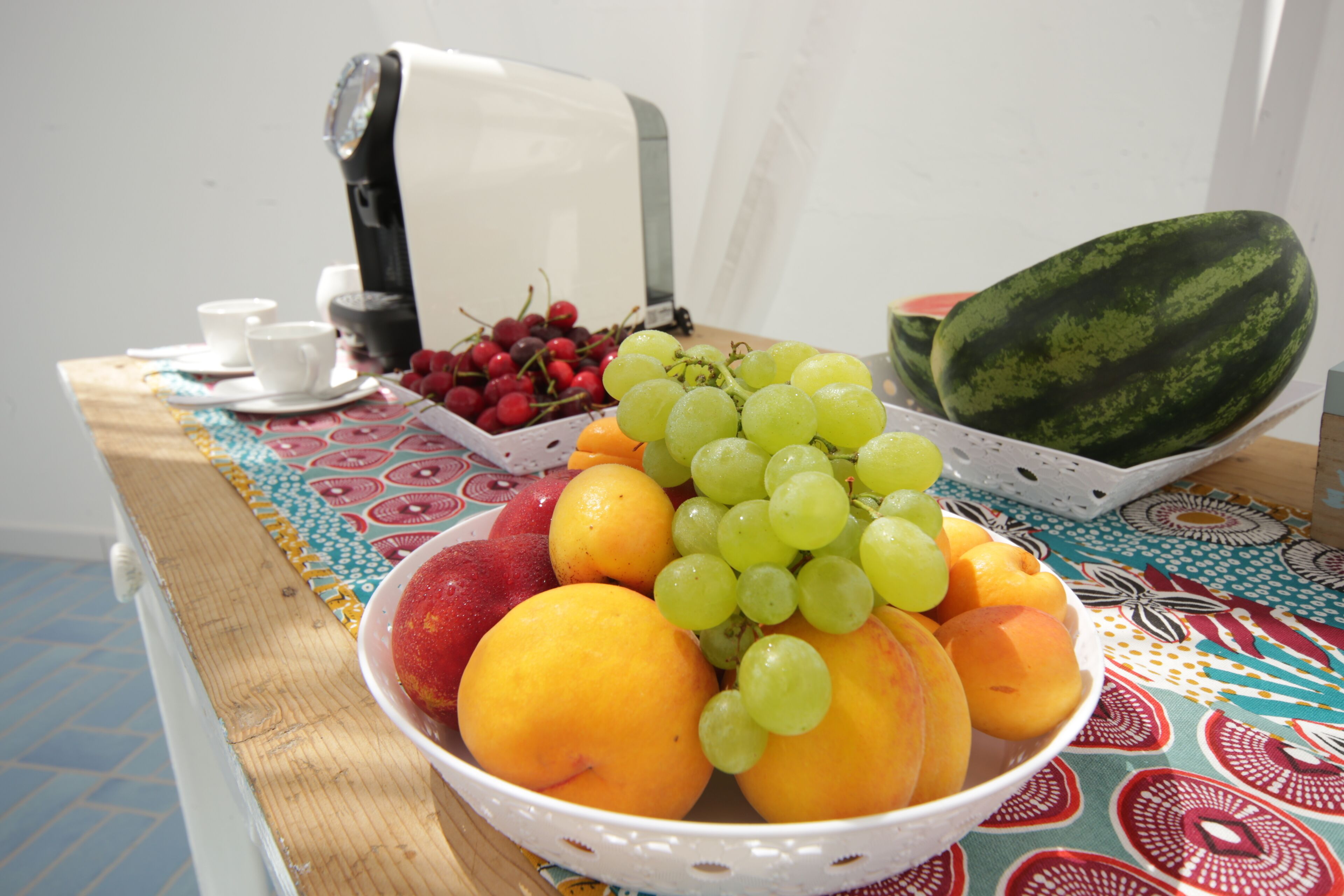 A table with a bowl of fruit including grapes, oranges, and a watermelon, and a coffee maker.