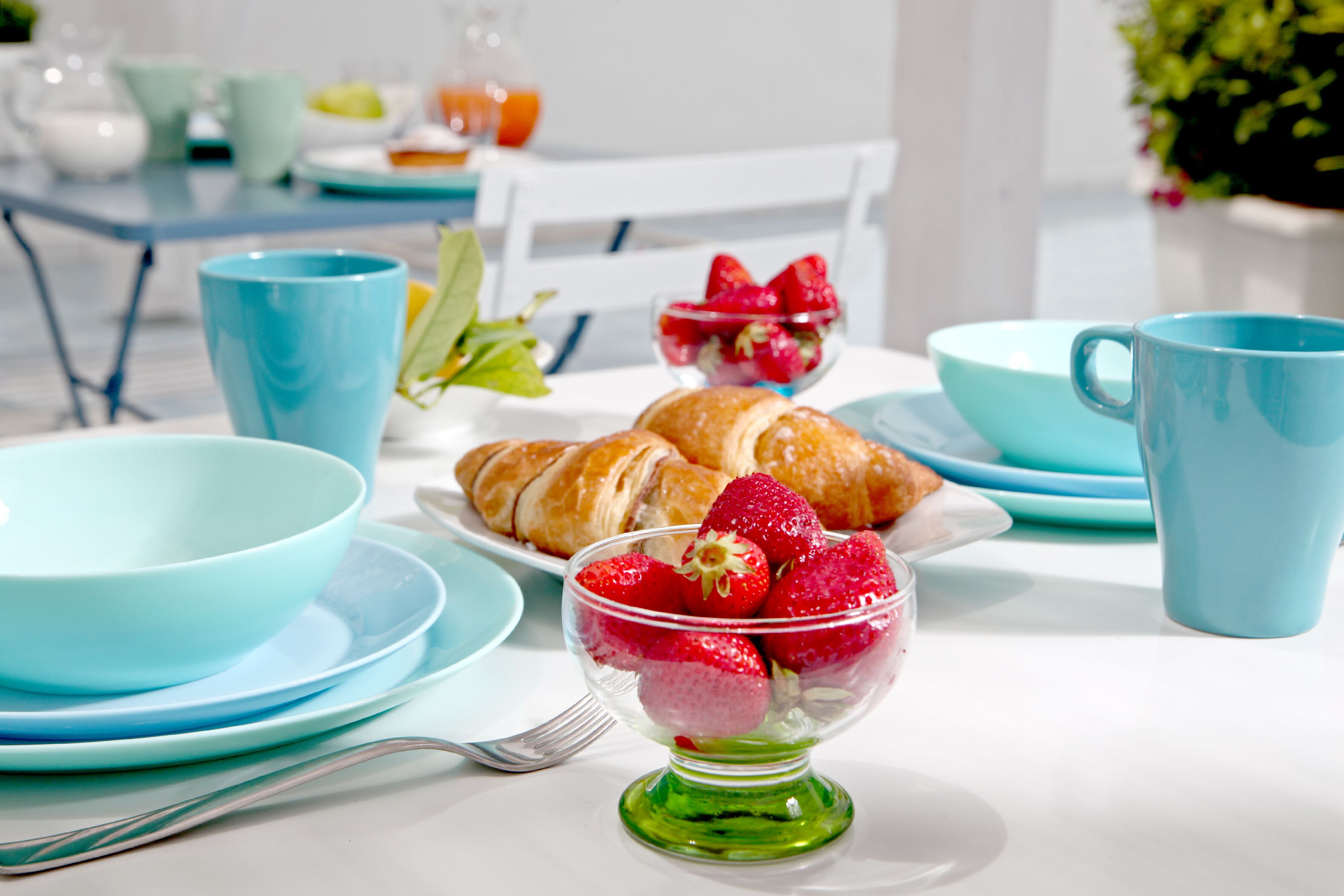 Un desayuno con una fuente de croissants, un bol de fresas y tazas y fuentes azules sobre una mesa blanca.
