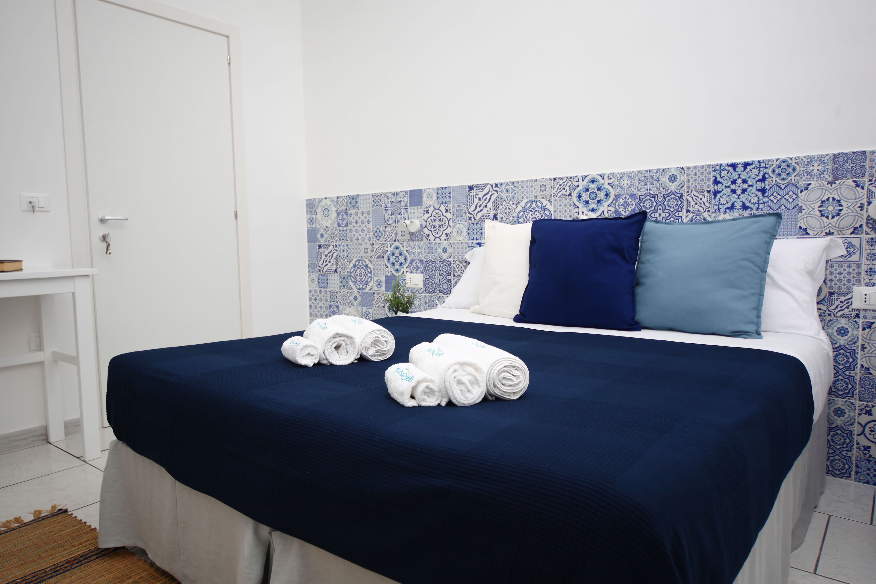 Cama bem arrumada com colcha azul escura, duas toalhas brancas enroladas e cabeceira estampada em azul e branco.