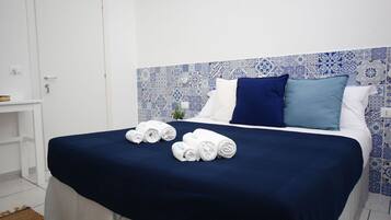Cama bem arrumada com colcha azul escura, duas toalhas brancas enroladas e cabeceira estampada em azul e branco.