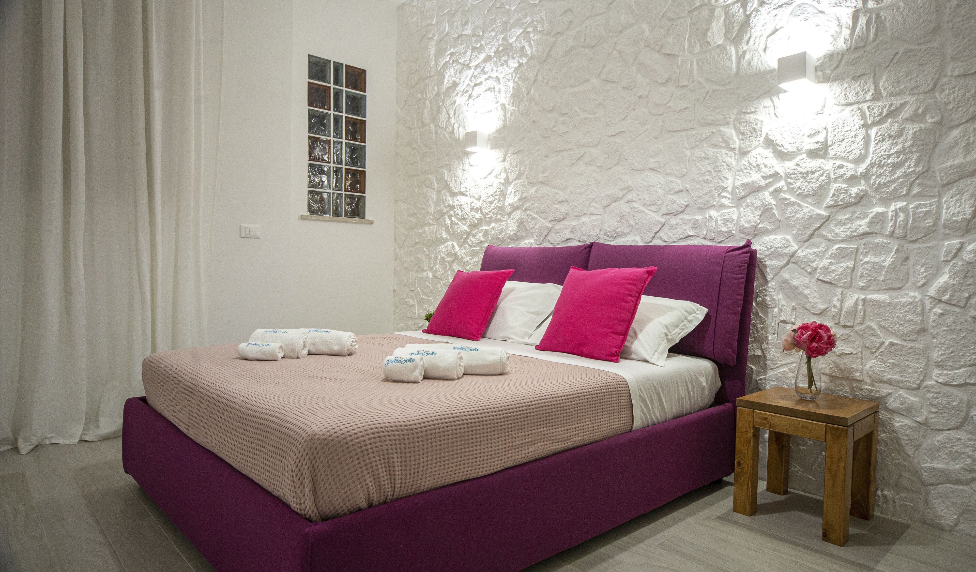A bedroom with a bed, pillows, a nightstand, a vase with a flower, and a window with a view of the outside.