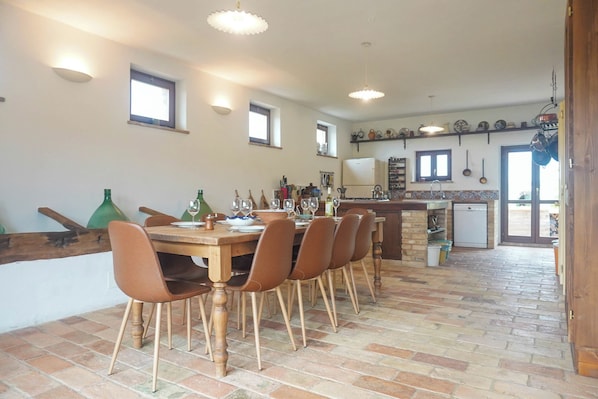 A dining area with a wooden table and chairs, a kitchen in the background, and a brick floor.