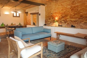 A cozy living room with a blue sofa, wooden chairs, a patterned rug, and a brick wall.