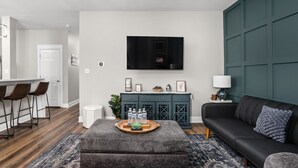 Una sala moderna con una pared de acento verde oscuro, un televisor de pantalla plana montado sobre un mueble de madera oscura, un sofá de cuero negro y un reposapiés gris con una bandeja de botellas de agua.