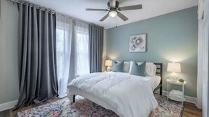 Un dormitorio con cama, ventilador de techo, mesita de noche con lámpara y alfombra floral.
