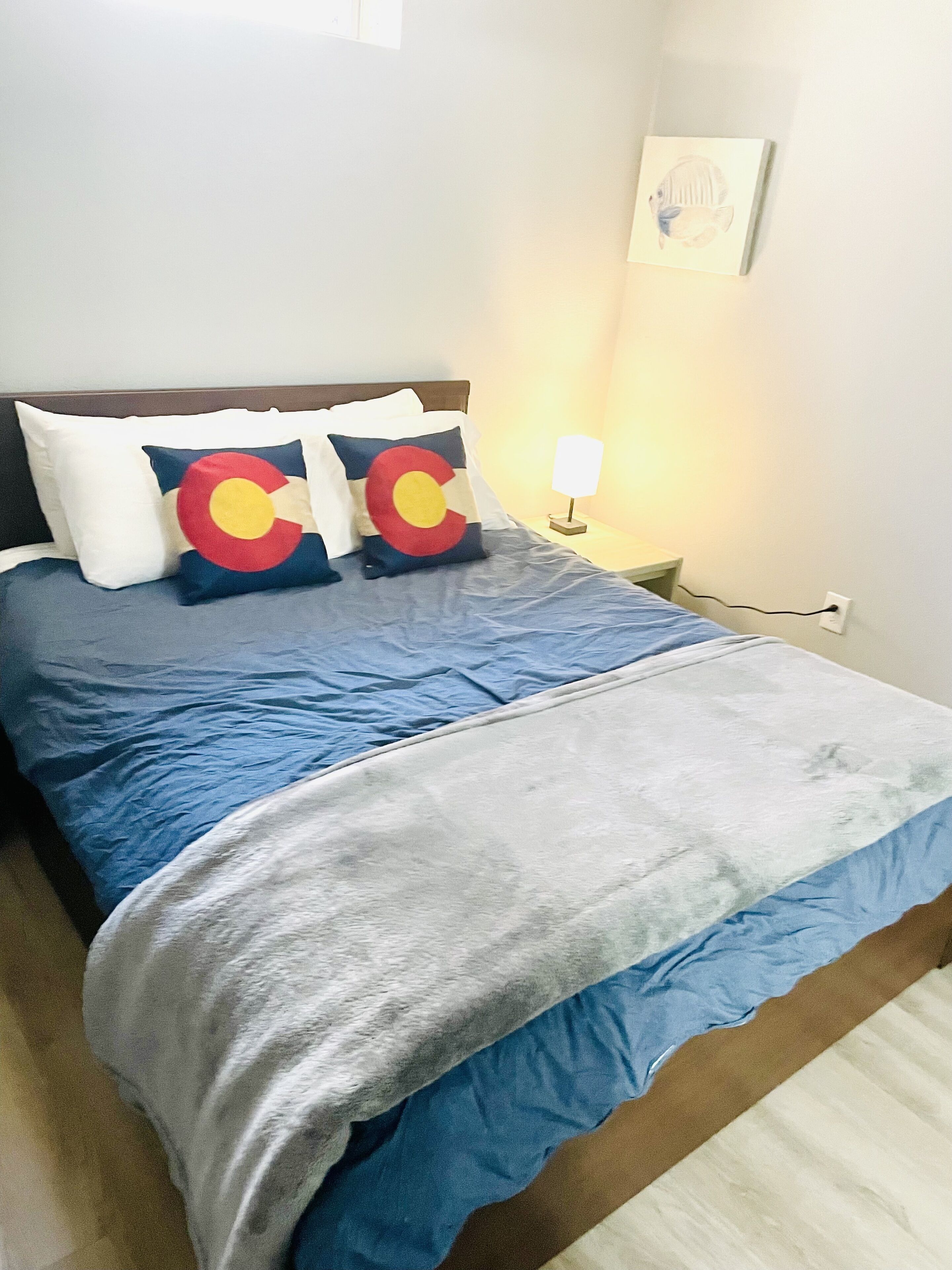A neatly made bed with blue and white bedding, two pillows with a red and yellow design, and a bedside lamp.