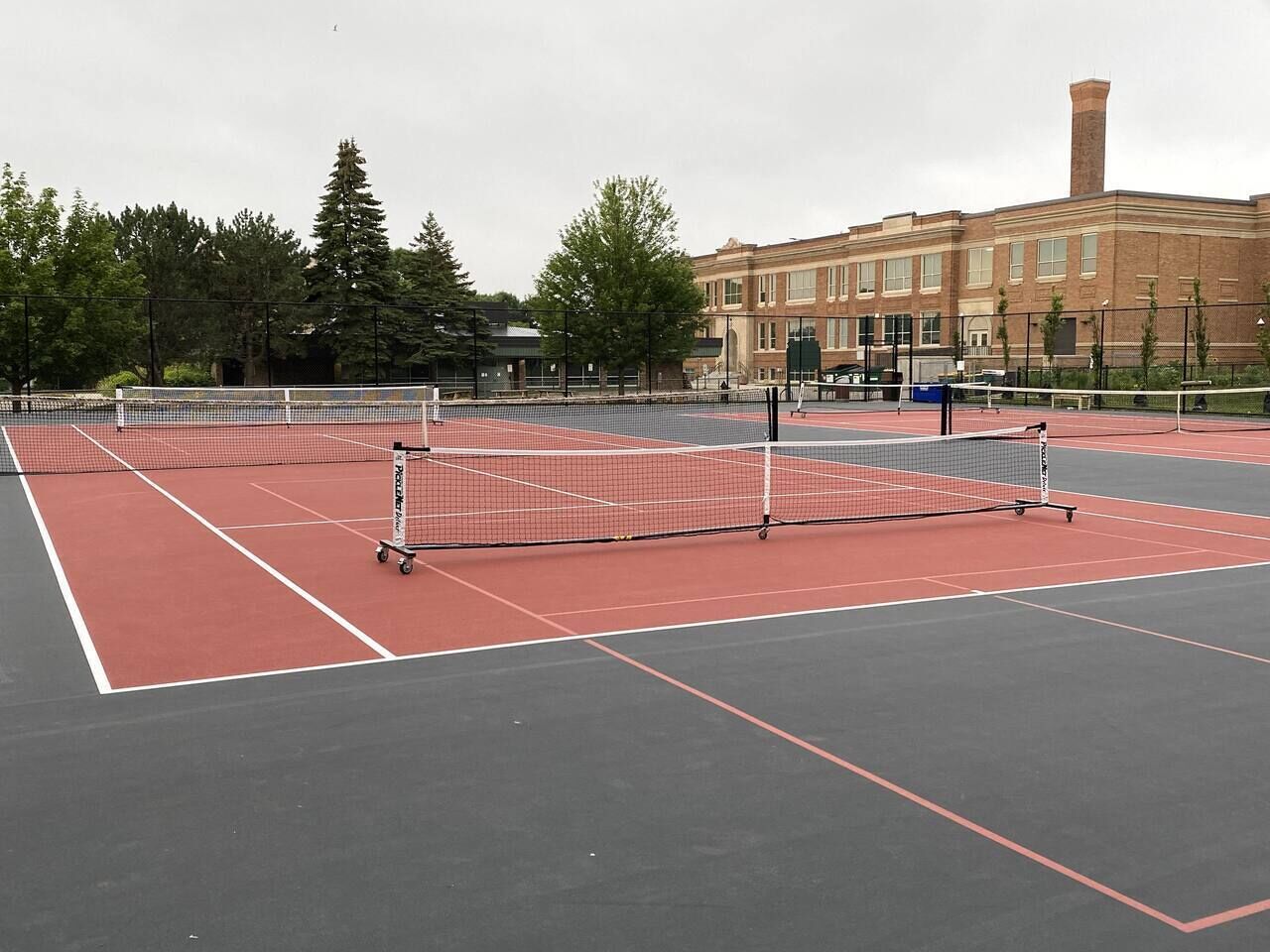 Ein Tennisplatz mit Netz, umgeben von Bäumen und einem Gebäude im Hintergrund.