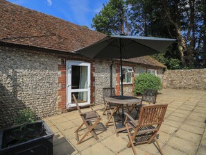 A stone building with a tiled roof, a patio with a table and chairs, and a large umbrella. - Byre Cottage 3 (Pulborough)