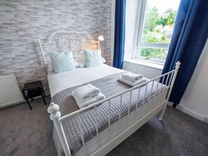 Een netjes opgemaakt bed met een wit metalen frame en grijze beddengoed, een klein nachtkastje en een raam met blauwe gordijnen.