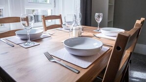 Ruokapöytä on katettu valkoisilla lautasilla, kulhoilla ja viinilaseilla. Pöydän ympärillä on puiset tuolit, ja taustalla näkyy ikkuna.