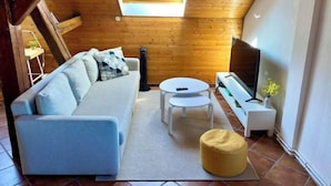 Ett mysigt rum i loftstil med ett träpanelat tak, en soffa, ett litet runt bord, en tv och ett fönster med utsikt över en byggnad.