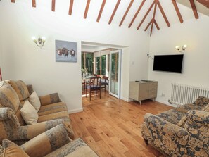 A spacious living area with wooden flooring, a sofa set, a television mounted on the wall, and a dining area with natural light.