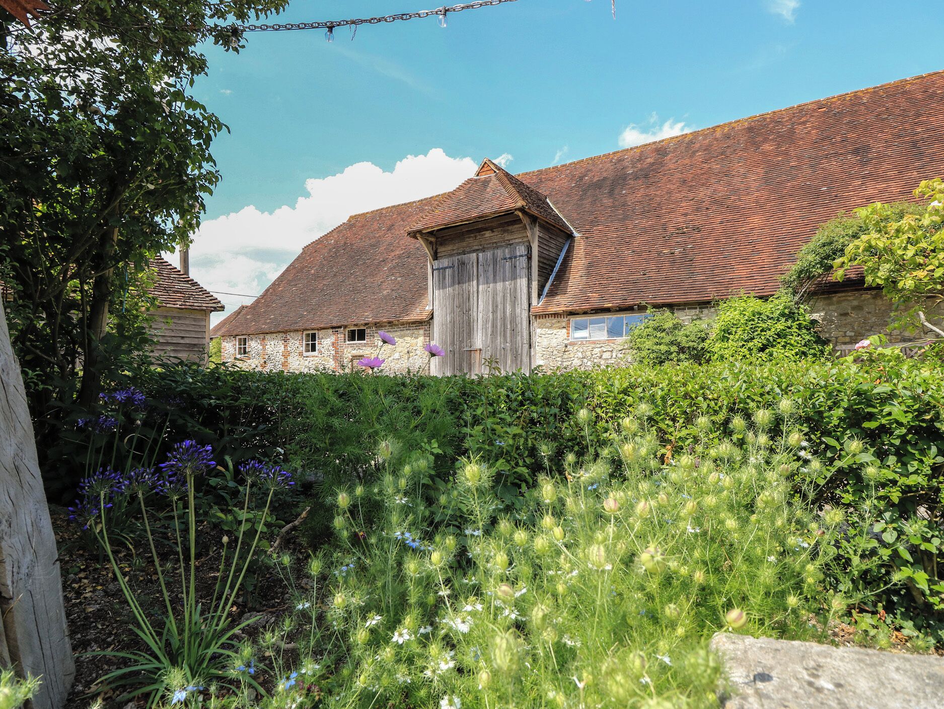 Une grange rustique, dotée d’un toit en pente, entourée d’une végétation luxuriante et de plantes en fleurs.