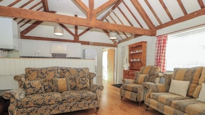 A spacious living area with wooden flooring, a floral-patterned sofa, a striped armchair, and a wooden cabinet with tea cups.