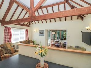 A room with a wooden ceiling, a sofa, a dining table with chairs, a TV, and a window with curtains. - Byre Cottage 4 (Pulborough)