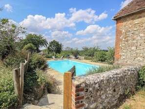 A swimming pool with a stone wall and a wooden fence, surrounded by greenery and a clear sky. - Byre Cottage 4 (Pulborough)