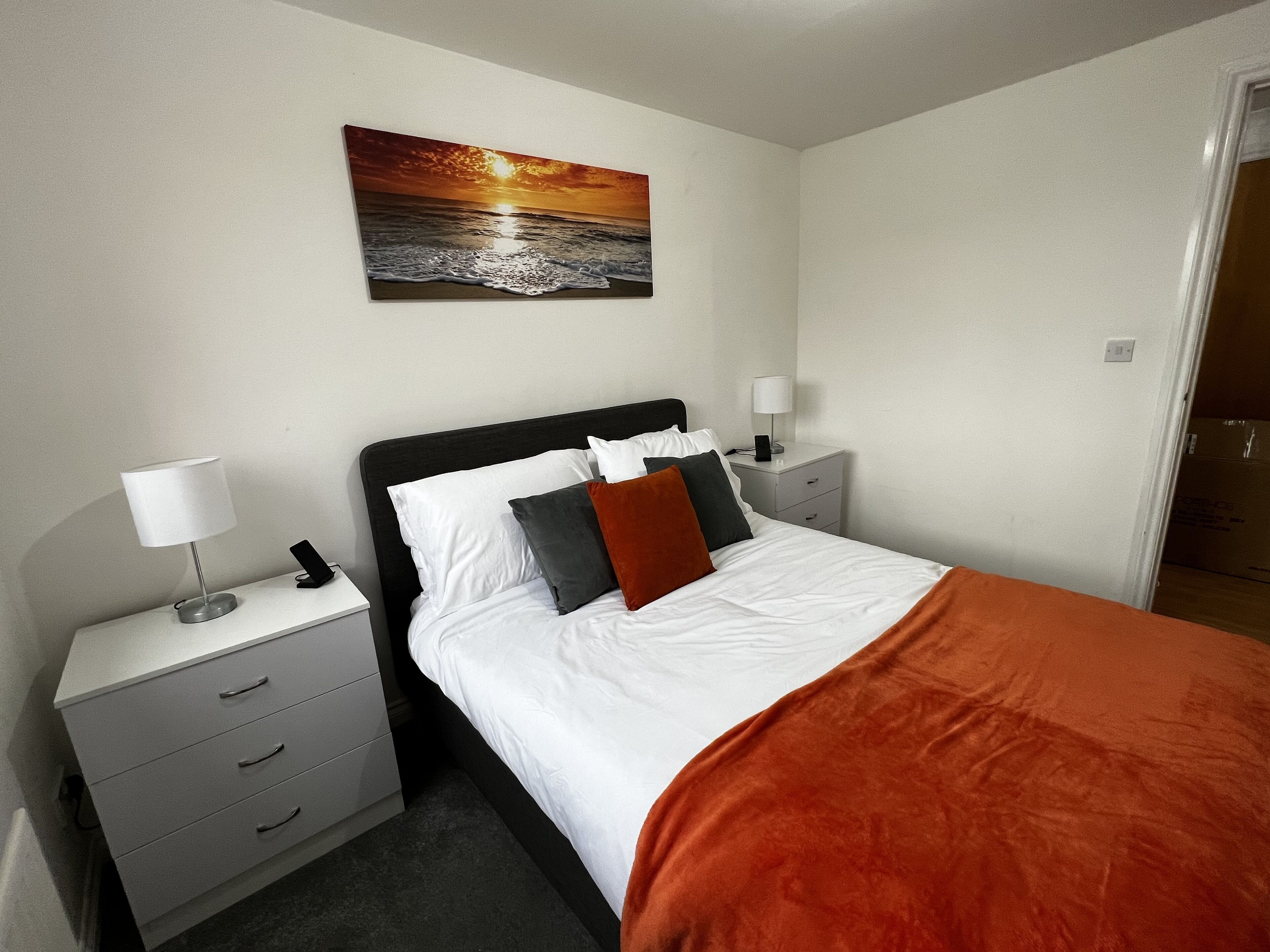 A neatly made bed with white and orange bedding, a black headboard, and a nightstand with a lamp and phone.