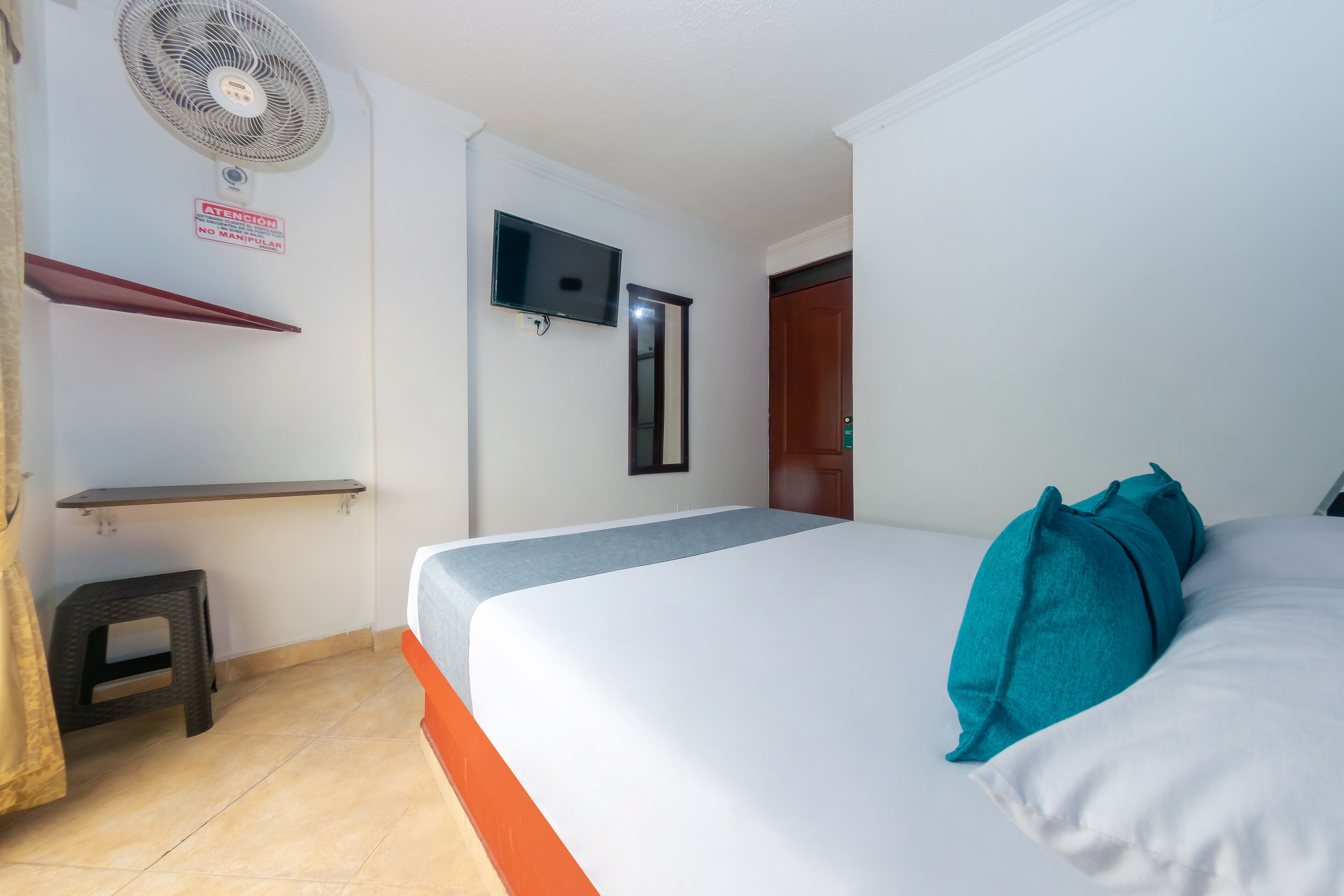 A hotel room with a bed, a TV mounted on the wall, a small stool, and a fan.