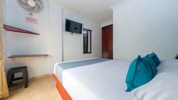 A hotel room with a bed, a TV mounted on the wall, a small stool, and a fan.