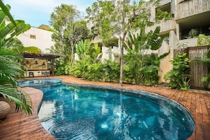 A swimming pool with a wooden deck, surrounded by lush greenery and a building with balconies.