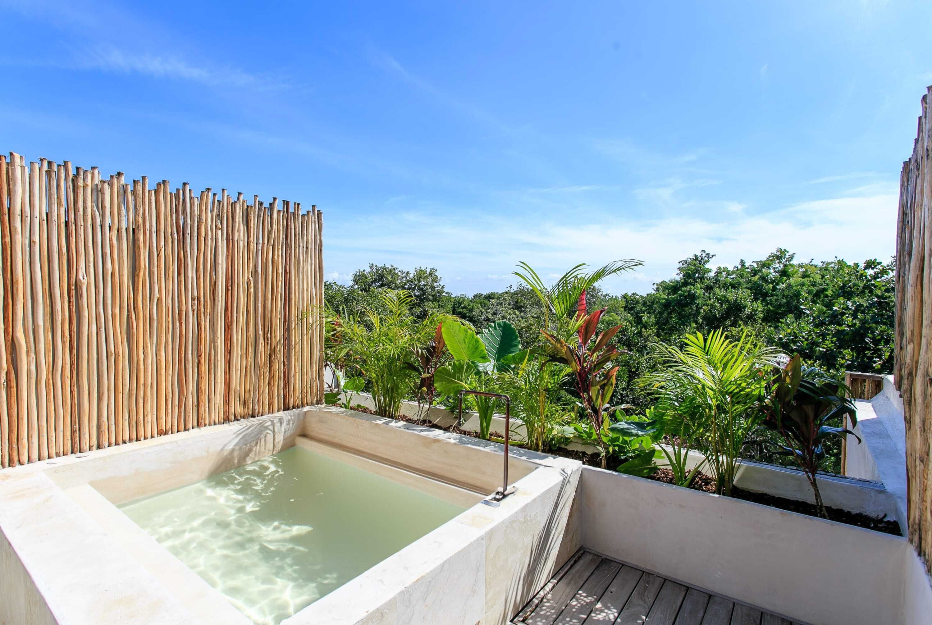 Un espace avec un bain à remous, délimité par une clôture en bambou et entouré d’une végétation luxuriante, sous un ciel dégagé.