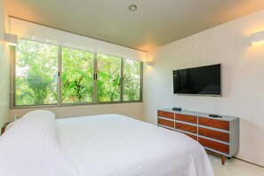 Une chambre avec une grande fenêtre, un lit, une télévision et une commode.