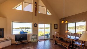 A spacious living room with a large window overlooking a body of water, a wooden dining table, and a television on a stand.