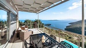A balcony with wicker furniture, a glass table, and a view of the sea and hills. - Dreamviewvillas (ΛΕΥΚΑΔΑ)
