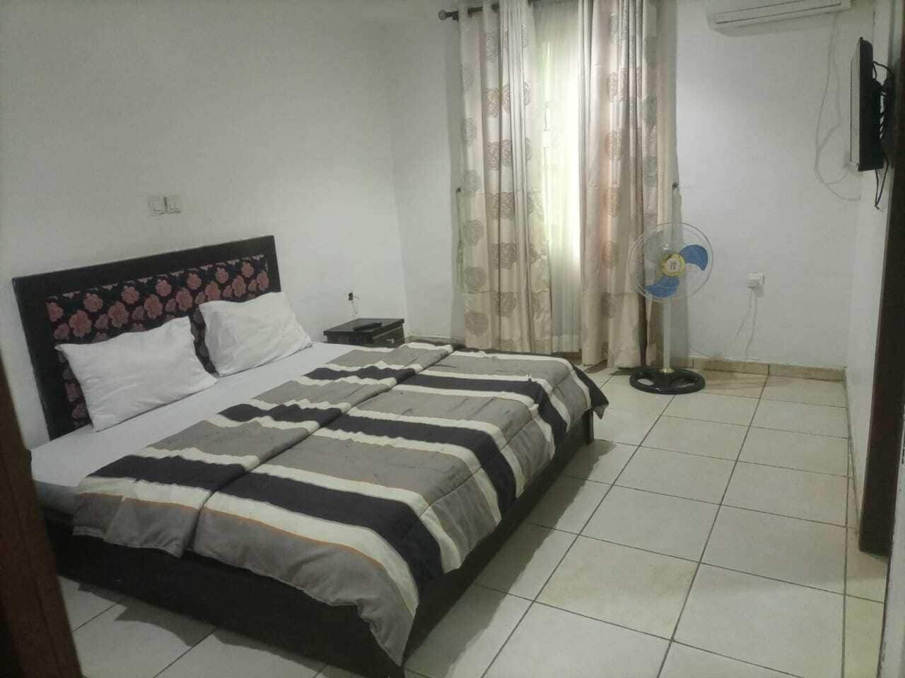 A bedroom with a double bed, a bedside table, a standing fan, and a curtain.