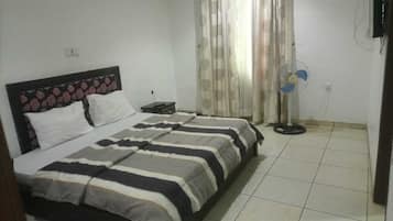 A bedroom with a double bed, a bedside table, a standing fan, and a curtain.