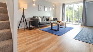 Un salon moderne avec un canapé gris, une table basse et un tapis bleu. Un escalier se trouve à gauche et une grande fenêtre à droite.