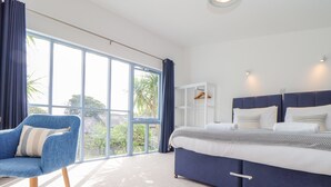 Une chambre d’hôtel moderne avec un grand lit, un fauteuil bleu et une vue sur l’extérieur grâce à de grandes fenêtres.