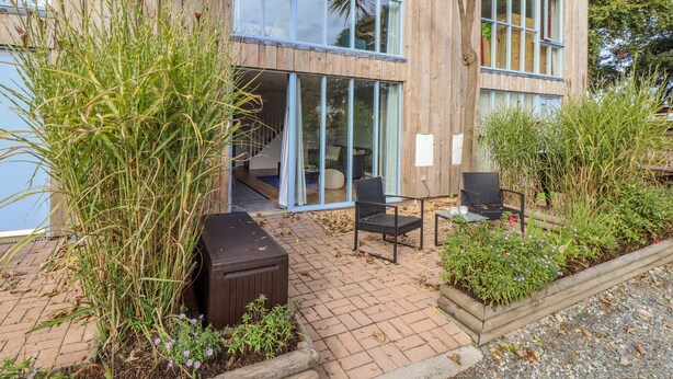 Un bâtiment moderne doté de grandes fenêtres, d’un patio aménagé avec du mobilier d’extérieur et d’une terrasse en bois.