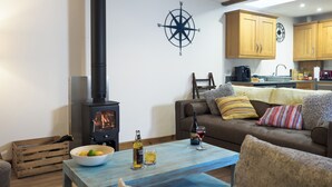 A cozy living room with a wood-burning stove, a coffee table with drinks, a couch, and a kitchen in the background. - Manacles (Falmouth)