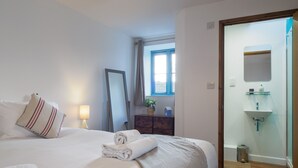 A hotel room with a bed, a bedside lamp, a mirror, a window, a dresser, a small plant, and a bathroom with a sink and mirror. - Manacles (Falmouth)