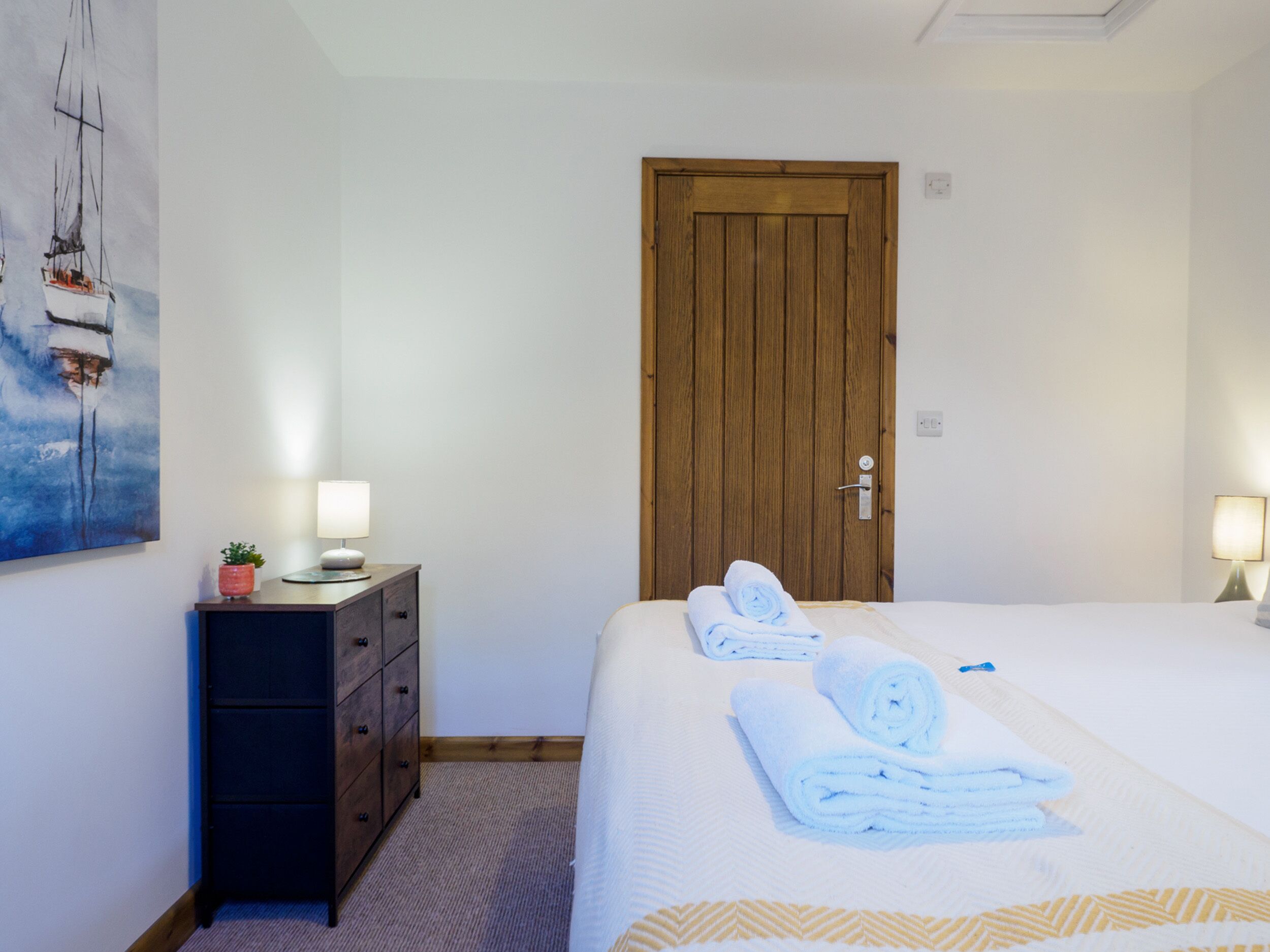 Habitación de hotel con puerta de madera, cama con ropa blanca, dos lámparas de noche, un tocador pequeño y un cuadro en la pared.