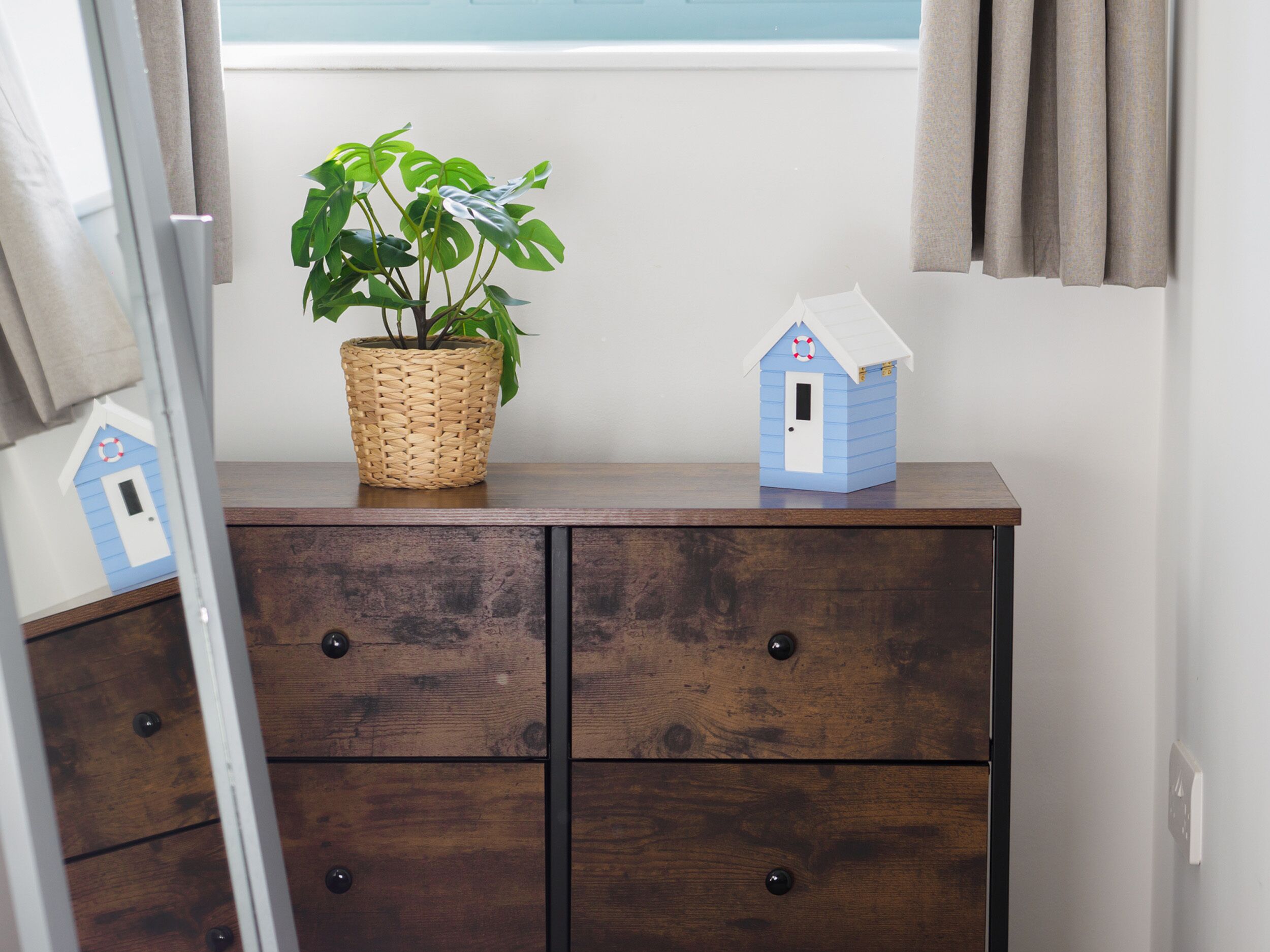 A wooden dresser with two drawers, a wicker basket with a plant, and a small blue house model on top.