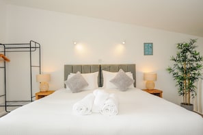 A neatly made bed with rolled towels, two bedside lamps, a wall-mounted shelf, a headboard, and a potted plant.
