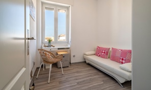A small, well-lit room with a white sofa, a wicker chair, a desk with a window, and a view of buildings outside.